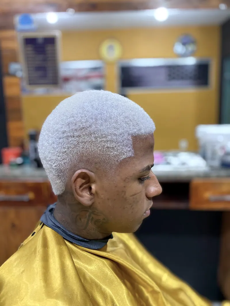 Corte platinado com o cabelo bem curto, de perfil, na barbearia de parede amarela